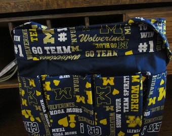 May include: A navy blue tote bag with multiple pockets, featuring a repeating pattern of yellow and white text and the letter "M". The text includes "Wolverines", "Go Team", and "#1".