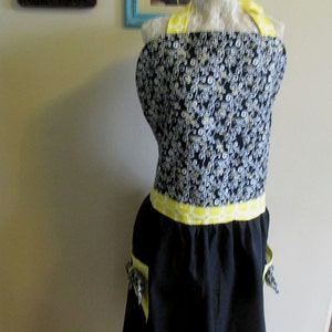 May include: Black and white patterned apron with yellow trim and pockets. The apron has a yellow and white geometric pattern around the waist.