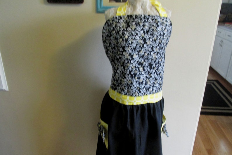 May include: Black and white patterned apron with yellow trim and pockets. The apron has a yellow and white geometric pattern around the waist.