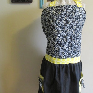 May include: Black apron with a yellow and white patterned waistband and two pockets. The apron has a black and white floral print on the top half.