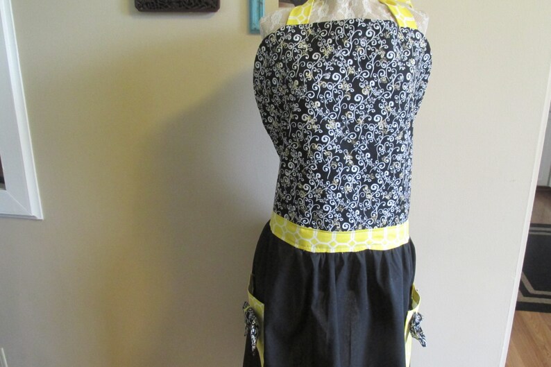 May include: Black apron with a yellow and white patterned waistband and two pockets. The apron has a black and white floral print on the top half.