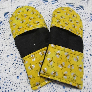 May include: A pair of yellow oven mitts with a black center and a pattern of bees flying around. The bees are silver and yellow.