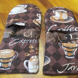 May include: A pair of brown and white oven mitts with a coffee theme. The mitts feature a checkered pattern and images of coffee cups and mugs. The text "Espresso", "Coffee", and "Java" is printed on the mitts.