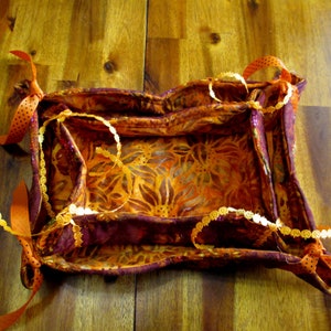 Könnte beinhalten: Ein handgefertigter Stoffkorb mit einem Blumenmuster in Orange- und Brauntönen. Der Korb ist mit einem dunkelburgunderfarbenen Stoff und orangefarbenem Band verziert. Eine goldfarbene Zierleiste ist ebenfalls vorhanden.