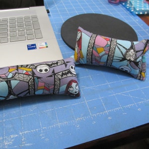 May include: Two purple and blue fabric wrist rests with a stained glass pattern and characters from the Nightmare Before Christmas. The wrist rests are on a blue cutting mat.