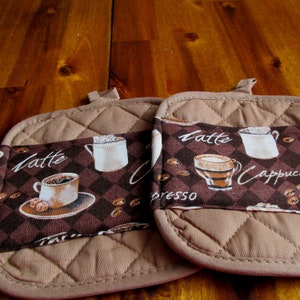 Könnte beinhalten: Zwei braune, gesteppte Topflappen mit Kaffeethema. Die Topflappen haben einen braunen Hintergrund mit einem weißen Rautenmuster. Die Texte "Latte" und "Cappuccino" sind auf den Topflappen gedruckt, zusammen mit Bildern von Kaffeetassen.