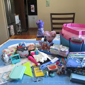 May include: A blue and pink fabric tote bag with the words "Quilt Tags" on the front. The bag is filled with various sewing supplies, including fabric scraps, thread spools, rulers, and a pair of white gloves. There is also a small purple fabric doll, a heart-shaped pincushion, and a bottle of liquid.