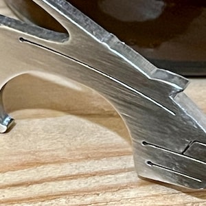 Puede incluir: Un abridor de botellas de metal plateado con un diseño único. El abridor tiene un mango curvo y una punta puntiaguda para abrir botellas.