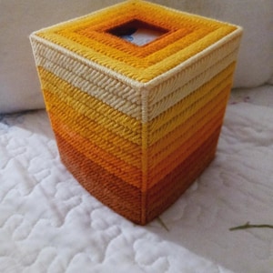 May include: A square tissue box cover made of plastic canvas with a gradient of orange, yellow, and white colors. The cover has a square opening for tissues.