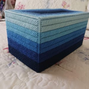 May include: A rectangular tissue box cover crafted from plastic canvas with a stitched yarn design. The exterior features horizontal stripes in a gradient of blue shades, transitioning from light sky blue at the top to a deep navy blue at the base.