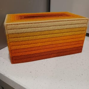 May include: A rectangular tissue box cover with a gradient design. The colors transition from light beige at the top to shades of orange and red at the bottom. The cover is made of a woven material.