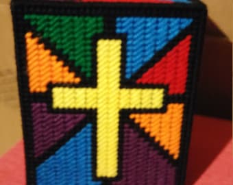 May include: A colorful tissue box cover with a yellow cross in the center. The cover is made of plastic canvas and features a stained glass design with various colors, including purple, blue, green, orange, and red.