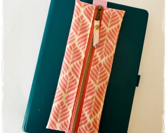 Könnte beinhalten: Ein Stifteetui mit rosa-weißem Muster und Reißverschluss. Das Etui ist mit einem rosa Gummiband an einem tealfarbenen Notizbuch befestigt.