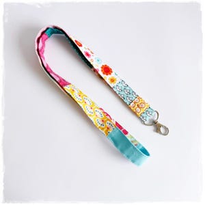 Könnte beinhalten: Ein farbenfrohes Schlüsselband mit Blumen- und Schmetterlingsmuster. Das Schlüsselband ist aus Stoff gefertigt und verfügt über einen Karabinerverschluss. Das Design umfasst Gelb-, Rosa-, Blau- und Orangetöne.