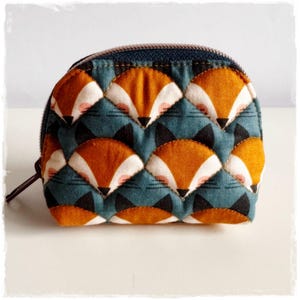 Könnte beinhalten: Ein kleines, halbkreisförmiges Reißverschlusstäschchen mit einem sich wiederholenden Steppmuster aus stilisierten Fuchsköpfen. Das Design zeigt orange, weiße und schwarze Fuchsköpfe auf einem gedeckten blauen Hintergrund mit dunklem Reißverschluss.