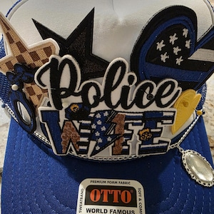 May include: A blue and white trucker hat with the word "Police" in script and the word "Wife" in block letters. The hat features various patches, including stars, a flag design, and a yellow emblem. The brim is blue, and the crown is white.