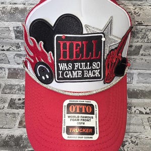 May include: A red and white trucker hat featuring a central rectangular patch with the text HELL WAS FULL SO I CAME BACK. The hat is decorated with a black heart, a silver star, red flames, and a black smiley face patch, with a silver trim.
