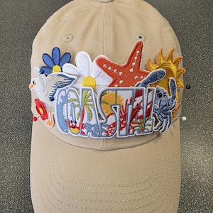 May include: Tan baseball cap with embroidered patches. Patches include a starfish, sun, flowers, and the word "COASTAL" in colorful letters. The cap is secured with pins.