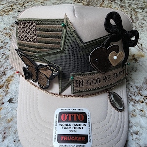 May include: A beige trucker hat with a mesh back and foam front. The hat is decorated with a brown and green American flag patch, a star, a butterfly, and the phrase "IN GOD WE TRUST". A black bow and two hearts are also attached.