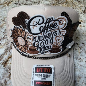 May include: Tan trucker hat with the phrase "Coffee is Always a Good Idea" embroidered in black and brown. Embellishments include a sunflower, hearts, and a butterfly. The hat has a black chain and a tag that says "OTTO".
