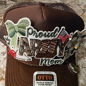 May include: A brown trucker hat with a mesh back and a curved brim. The hat features an embroidered patch with the words "Proud Army Mom", a camouflage design, and an American flag.