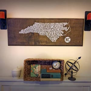 May include: A wooden sign with a white outline of the state of North Carolina, filled with the names of cities and towns. The sign is hanging on a wall above a shelf with a basket, books, and a clock. The sign also has the letters "NC" in a circle.