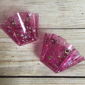 May include: Two translucent pink cupcake-shaped holders featuring a glittery finish with embedded holographic star-shaped sequins. Each holder has a scalloped top edge and two silver-toned metal eyelets positioned near the upper rim for attachment.