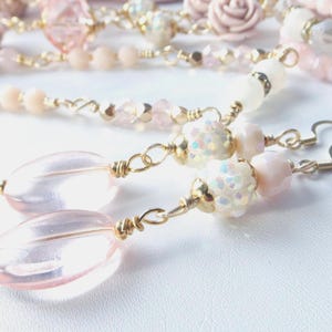 May include: Dangle earrings featuring pale pink oval beads, white iridescent textured beads, and gold-toned wire accents. The earrings are connected to a matching beaded chain necklace with additional pink beads, faceted crystals, and a small rose-shaped charm.