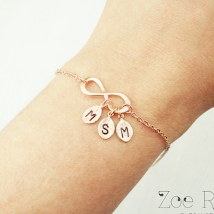 May include: Rose gold infinity bracelet with three leaf charms, each bearing an initial. The bracelet is delicate and elegant, ideal for a personalized gift. The chain is fine, and the infinity symbol is a prominent feature.