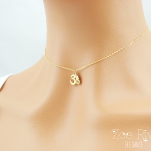May include: A delicate gold-colored necklace featuring an Om symbol pendant. The pendant is suspended from a fine chain, creating a minimalist and elegant design. The necklace is worn around the neck.