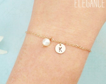 May include: A delicate gold bracelet with a small pearl charm and a round charm engraved with the letter "K". The bracelet is displayed on a wrist, with the text "Zoe River Elegance" in the background.