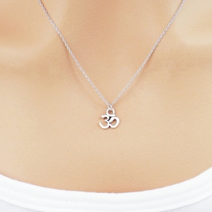 May include: A silver-colored necklace with a small Om symbol pendant. The necklace is displayed on a person wearing a white shirt. The brand name "Zoe River ELEGANCE" is visible in the upper left corner.