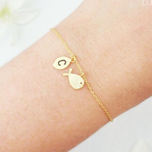 May include: A gold bracelet with a fine chain, showcasing a fish charm and a leaf-shaped charm with the letter "C". The bracelet is worn on a wrist, with the text "Zoe River Elegance" in the background.