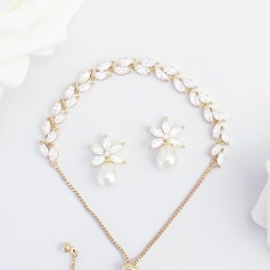 May include: A gold-coloured bracelet and matching earrings set. The bracelet features a floral design with clear crystals. The earrings have a flower shape with a dangling pearl. The set is displayed on a white surface with white roses.