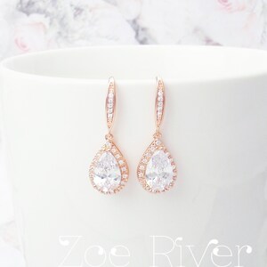 Peut inclure: Boucles d'oreilles en forme de goutte d'eau en or rose, avec un halo de petits cristaux transparents autour d'un grand cristal clair en forme de poire. Les boucles d'oreilles sont pr&eacute;sent&eacute;es sur une surface blanche. Le texte "Zoe River Elegance" est visible.