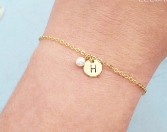 May include: Gold-colored chain bracelet with a small round charm engraved with the letter "H" and a small white pearl. The bracelet is delicate and elegant, perfect for everyday wear. The brand name "Zoe River Elegance" is visible.