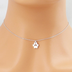 May include: A silver-coloured necklace with a paw print charm. The charm is a detailed cutout of a paw, hanging from a fine chain. The necklace is displayed on a person's neck. The brand name "Zoe River Elegance" is visible in the image.