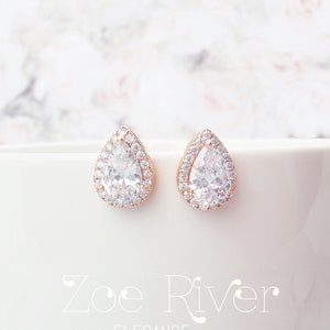 May include: Rose gold teardrop stud earrings. Each earring has a large, clear, pear-shaped central stone, encircled by smaller, clear stones. The earrings are displayed on a white surface with the text "Zoe River Elegance".