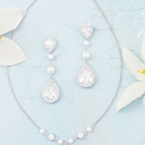 May include: A silver necklace and matching earrings set. The jewellery features pear-shaped, clear crystal pendants surrounded by smaller crystals. The earrings and necklace also include small, round pearls. The background is a light blue with white flowers.