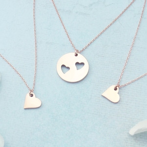 May include: Rose gold necklaces with heart pendants. One necklace features a round pendant with two heart cutouts. Two other necklaces have solid heart pendants. The necklaces are displayed on a light blue surface with white flowers.