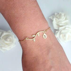 May include: A gold bracelet with a vine design and clear crystal details. The bracelet has a charm with the letter "S". The bracelet is worn on a wrist, with white roses in the background.