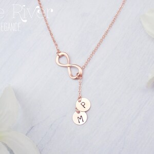 Könnte beinhalten: Halskette aus Roségold mit einem Unendlichkeitssymbol und zwei kleinen, runden Anhängern. Die Anhänger sind mit den Buchstaben "P" und "M" graviert. Die Halskette wird vor einem weichen, weißen Hintergrund mit floralen Akzenten präsentiert. Der Text "Zoe River Elegance" befindet sich oben links.