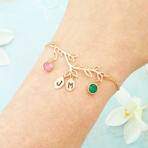 May include: A gold-coloured bracelet with a branch design, leaves, and hanging charms. It features a pink gemstone, a green gemstone, and two oval charms with the letters "J" and "M". The bracelet is worn on a wrist.