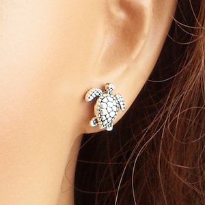 May include: A silver turtle stud earring. The earring is a detailed miniature of a turtle, with a textured shell and flippers. The earring is shown on a mannequin's ear, highlighting its size and design.