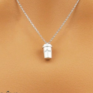 May include: A silver necklace with a miniature coffee cup pendant. The word "coffee" is written on the cup. The necklace is displayed against a white top.