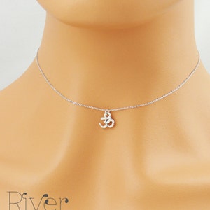 May include: A silver necklace with an Om symbol pendant. The pendant hangs from a fine chain, displayed around the neck. The image is a close-up, emphasising the necklace's details and the chain's subtle gleam. The necklace is a piece of jewellery.