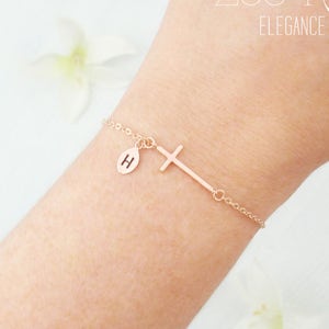 May include: Rose gold bracelet with a sideways cross and a small circular charm with the letter "H". The bracelet is attached to a fine chain. The text "Zoe River Elegance" is in the upper right corner.