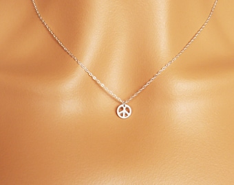 May include: A delicate silver necklace featuring a small peace symbol pendant. The pendant is a cut-out design, and the chain is fine and elegant. The necklace is shown on a person wearing a white shirt.