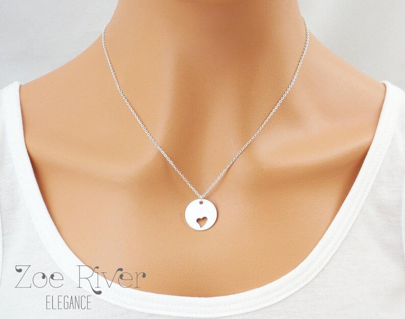 May include: A silver necklace featuring a delicate chain and a circular pendant with a heart-shaped cutout. The necklace is worn against a white top. The brand name "Zoe River Elegance" is visible at the bottom of the image.
