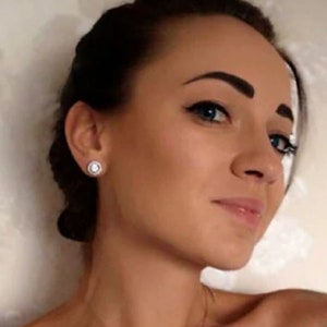 May include: A close-up shot of a person wearing a pair of round, silver stud earrings with a white, circular center stone and a halo of smaller stones. The earrings are set against the person's skin and dark hair.